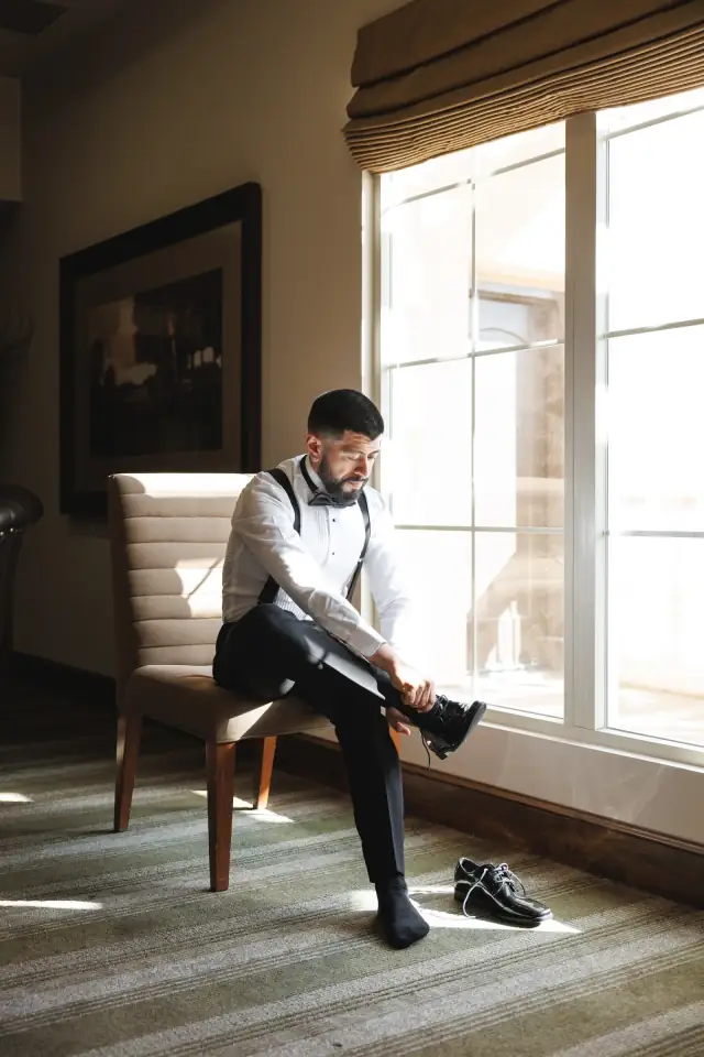 Groom sitting on a chair by a window putting on black dress shoes in a softly lit room during wedding preparations.