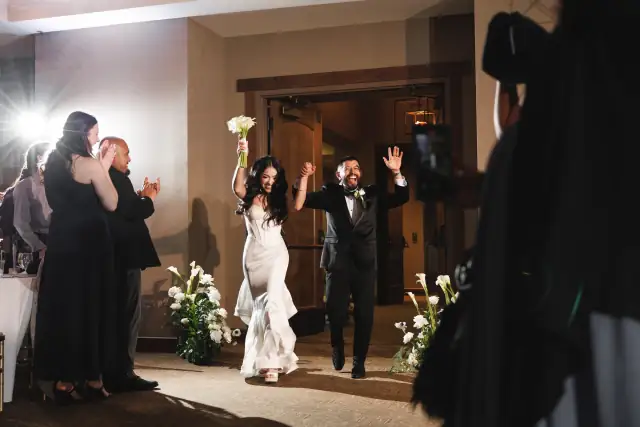 Bride and groom joyfully entering wedding reception hall holding hands, bride raising bouquet, guests clapping indoors.