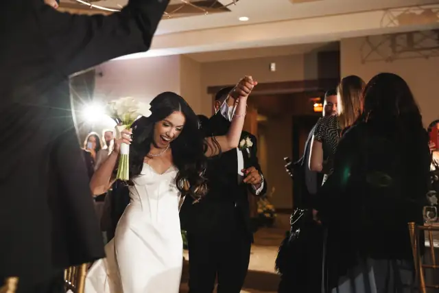 Bride in a white dress joyfully holding a bouquet and raising hands with the groom in a dark suit during a wedding celebration indoors.