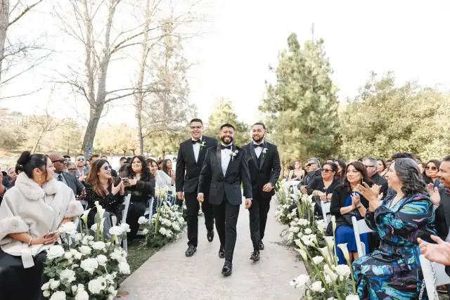 Groom and groomsmen walking down outdoor aisle lined with white flowers while guests applaud at Grace & Oscar's wedding.