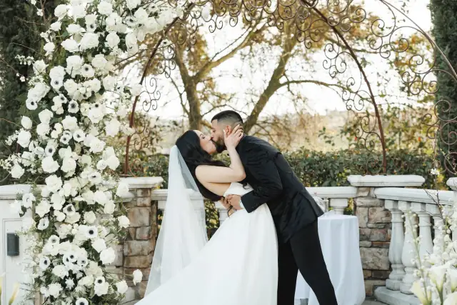 Bride and groom sharing a kiss under a floral arch at an outdoor wedding ceremony.