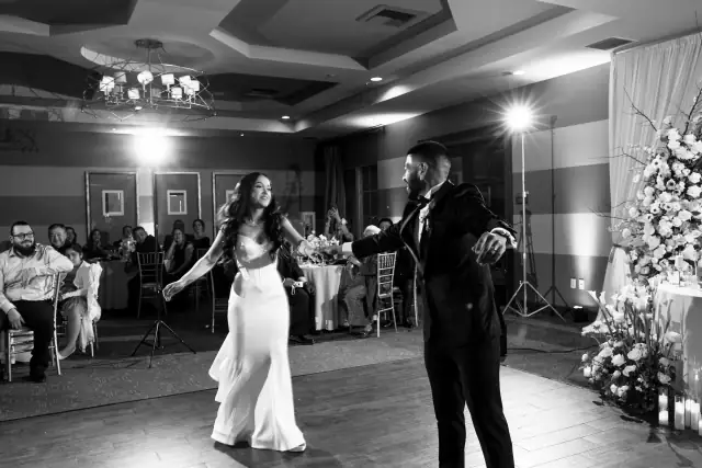 Bride and groom dancing at their indoor wedding reception surrounded by seated guests and floral decorations.