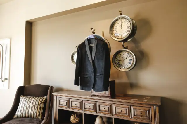 Black tuxedo jacket hanging on a vintage clock fixture above a wooden cabinet in a cozy room with a cushioned chair, wedding preparation setting.