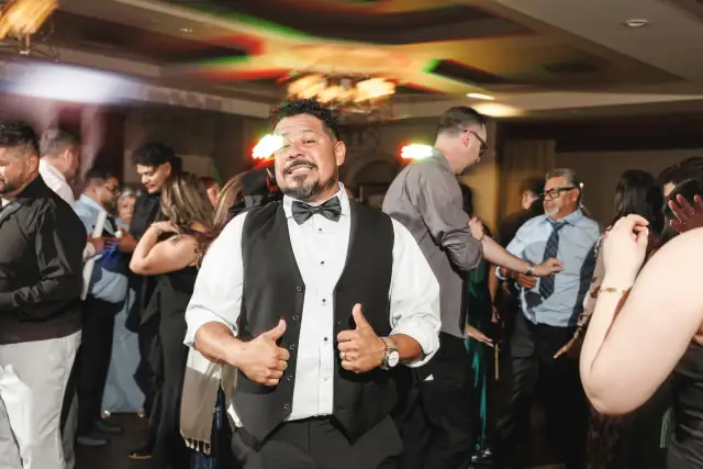 Man in tuxedo vest and bow tie giving thumbs up while guests dance at indoor wedding reception for Grace & Oscar by Zatonsky Moments.