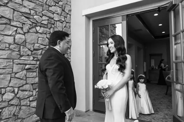 Bride in wedding dress holding bouquet smiling at man in suit outside stone building entrance with two flower girls and another woman inside.