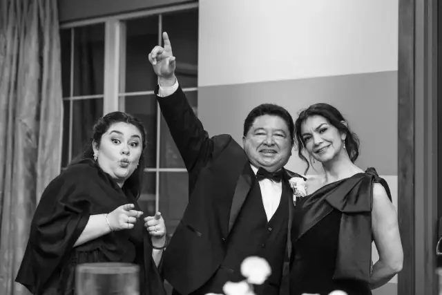 Black and white photo of three wedding guests posing happily indoors, with a man in a tuxedo and two women in elegant dresses at a formal event.