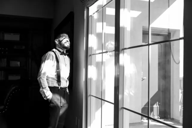 Groom in tuxedo with suspenders standing indoors by a window with sunlight and reflection, captured in a black and white wedding photo.