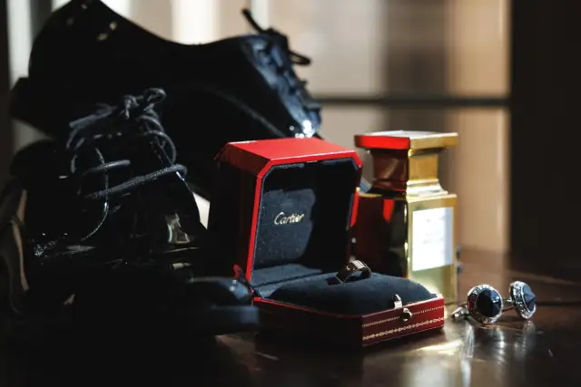 Close-up of groom's black patent leather shoes, Cartier wedding ring in red box, gold perfume bottle, and silver cufflinks on wooden surface.