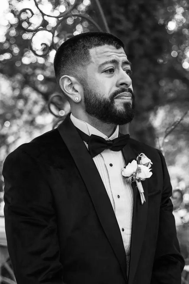 Groom in a tuxedo with a boutonniere, standing outdoors with an emotional expression during Grace and Oscar's wedding.